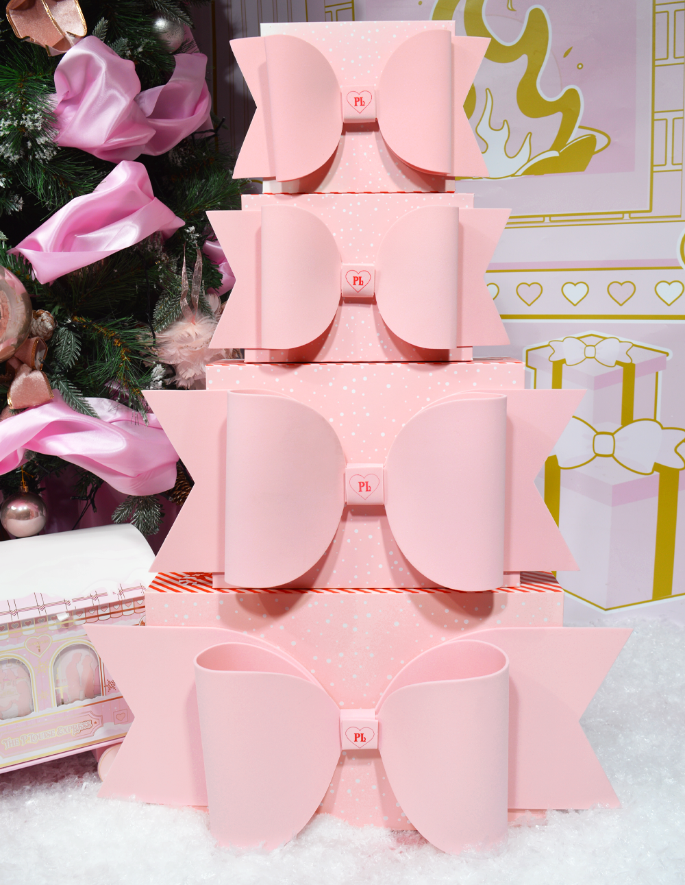 Stabel med rosa P.Louise-gaveesker med sløyfer utstilt mot et bakteppe med juletema med dekorasjoner.