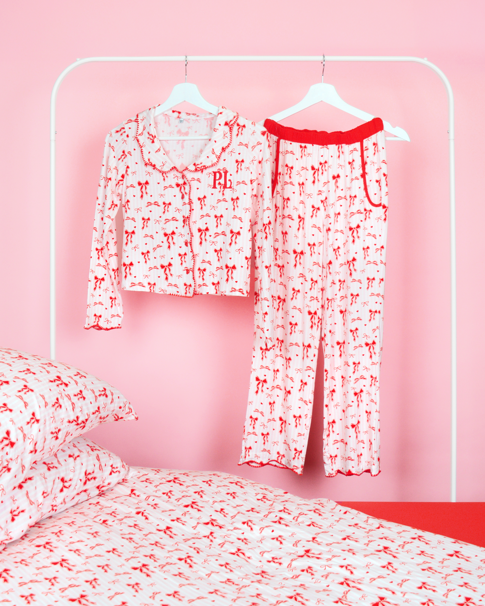 Mädchen-Pyjama-Set mit weihnachtlicher Schleife von P.Louise mit durchgehend rotem Schleifenmuster auf rosa und weißen Streifen, die an Kleiderbügeln in einem rosafarbenen Schlafzimmer mit passender Bettwäsche hängen
