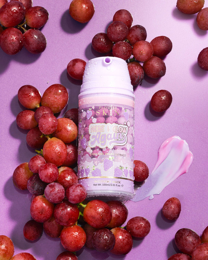 Paarse fles P. Louise Here I Glow Again Grape Recovery Mask omgeven door druiven op een paarse achtergrond