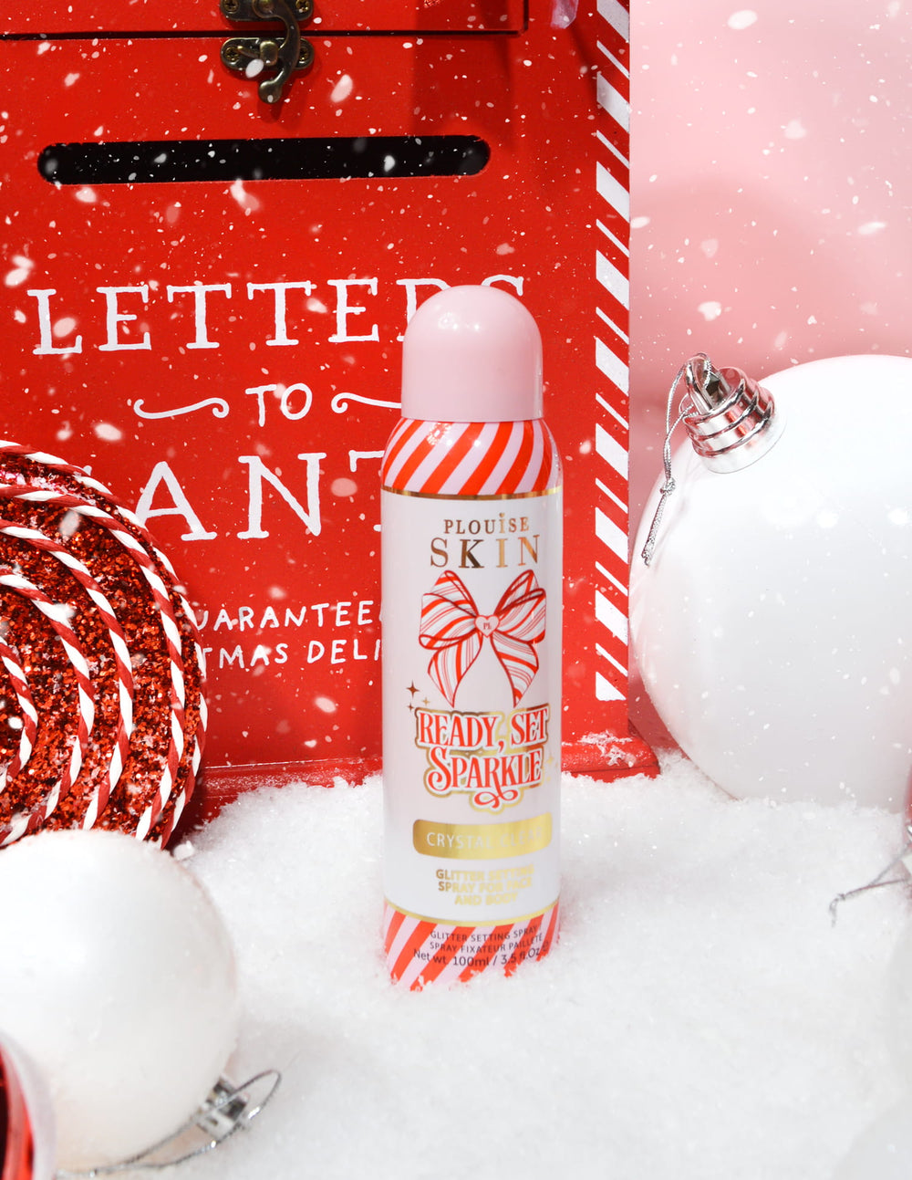 Une bouteille de P. Louise Ready Set Sparkle Christmas Glitter Setting Spray entourée de décorations festives comme de la neige, des boules et une boîte aux lettres de Noël