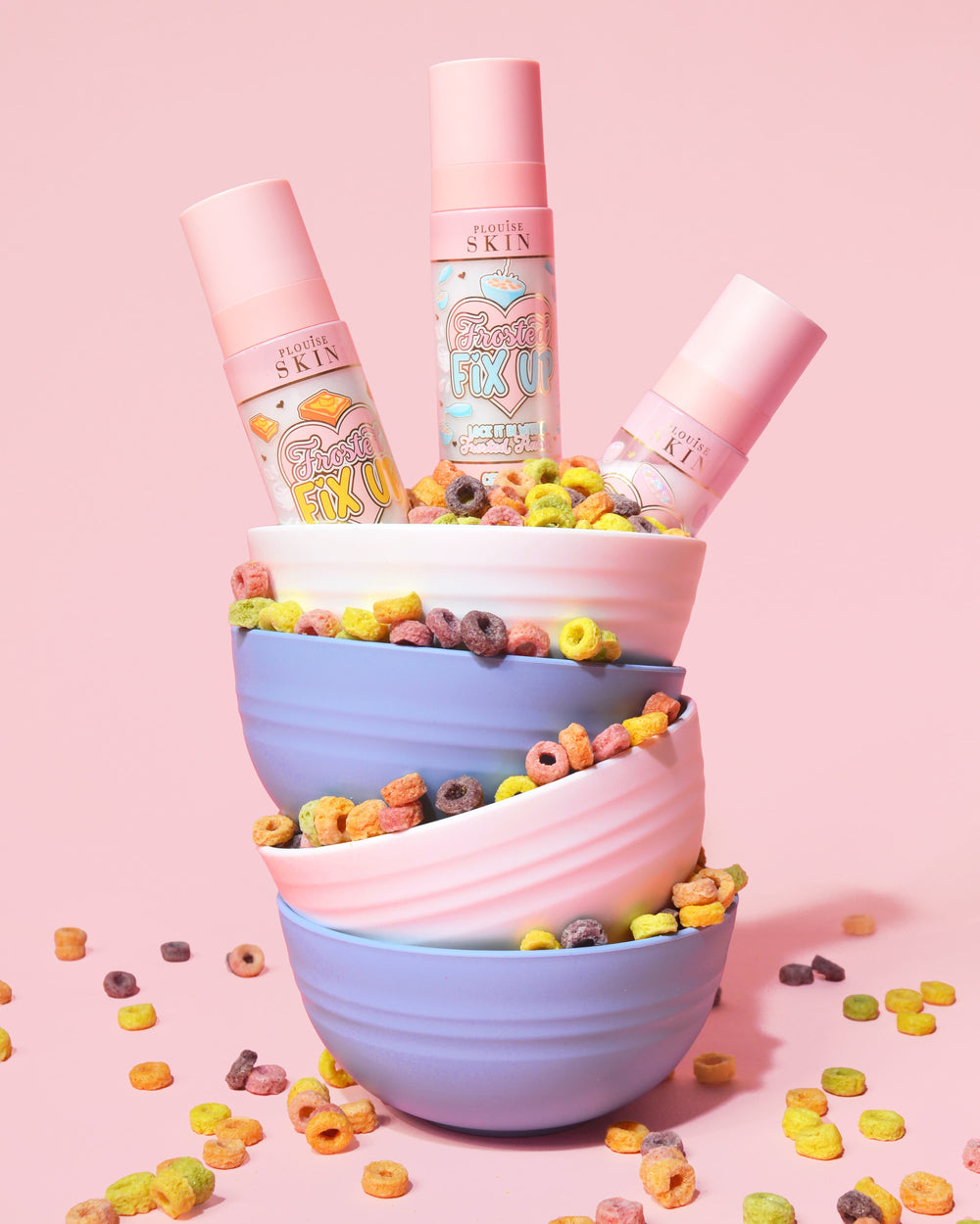Hög med färgglada skålar fyllda med flingor som häller ut och tre flaskor med P. Louise Frosted Fix Uo Blurring Setting Spray i den översta skålen på en rosa bakgrund