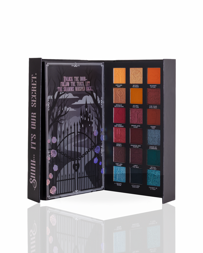 An open page of a halloween eyeshadow palette showing 18 colour pans in a gothic-themed box with a haunted house illustration on the inner lid. The open palette displays a mix of matte, shimmer, and glitter shades in orange, red, brown, blue, and green tones.