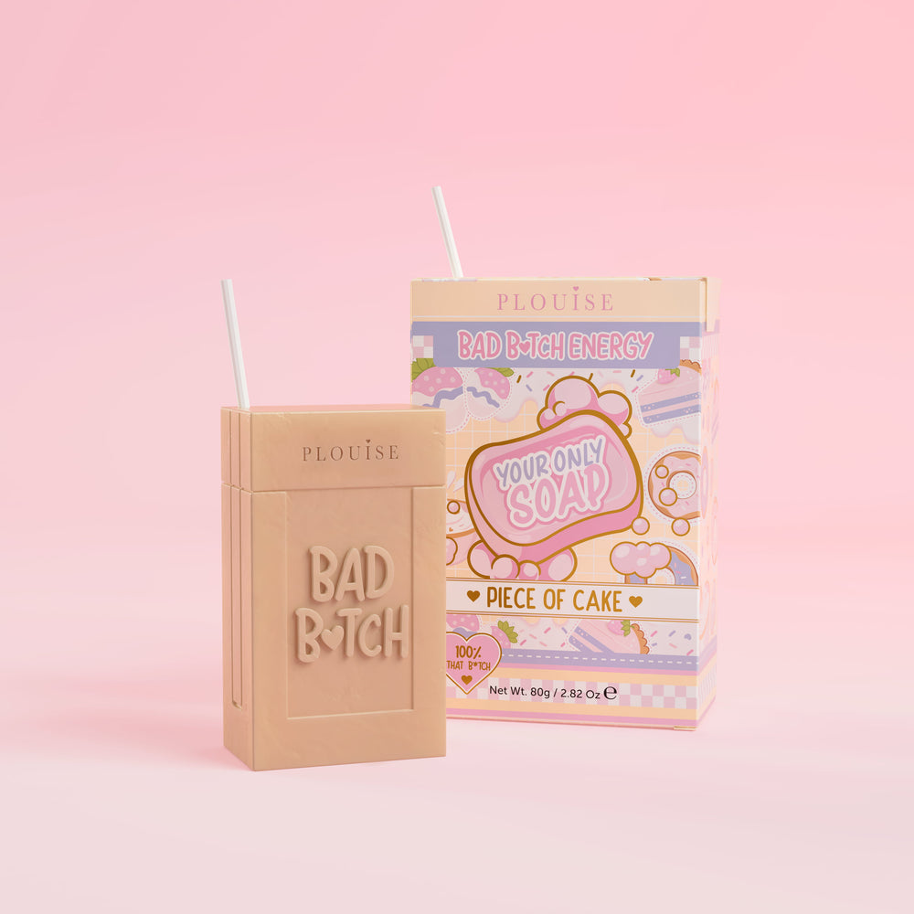 Et beige såpestykke med 'Bad Bitch' gravert på, ledsaget av en fargerik juicebokspakke med 'Piece of Cake' merket på, presentert på en rosa bakgrunn.