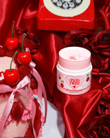 En beholder med P.Louise Plot Twist Make It Melt Makeup Cleansing Balm vist på en rød stofoverflade med dekorative elementer, herunder et kirsebær og en vintage rød telefon i baggrunden.