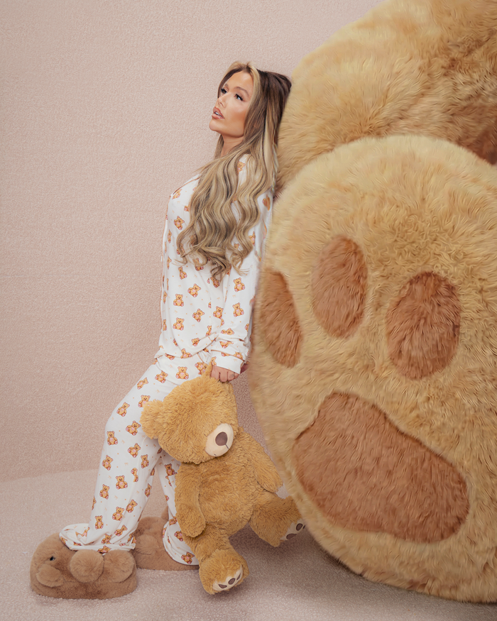 Kvinna i nallepyjamas som håller en nallebjörn bredvid en stor plysch björntass på en beige bakgrund