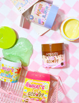 Une gamme de contenants de gommage corporels extensibles et colorés portant le nom du produit « What's the Scoop ? » affiché en évidence. Les conteneurs sont disposés sur une surface avec un fond à carreaux roses, et des graphiques et des textes ludiques et colorés sont associés au produit.