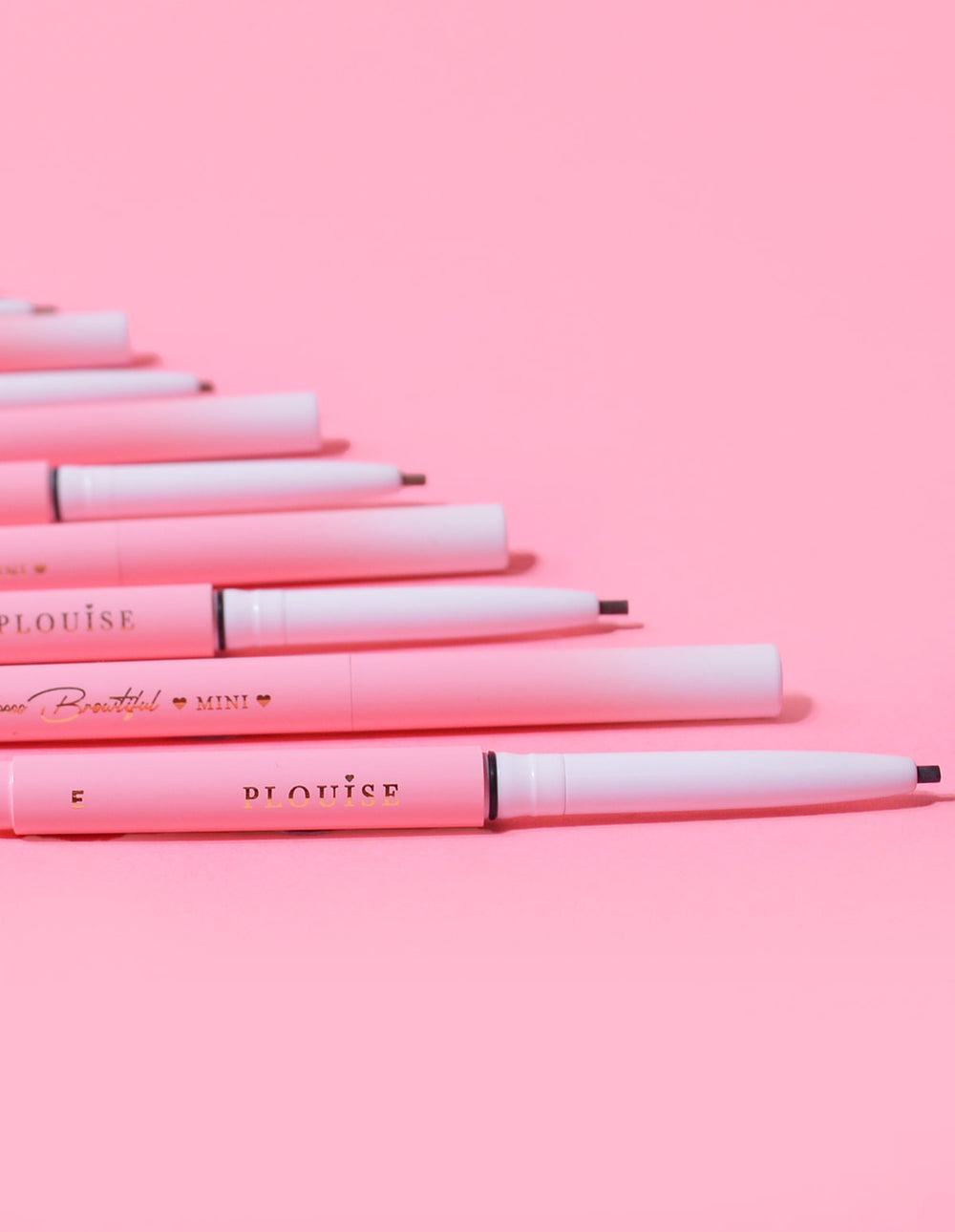 Una colección de mini lápices de cejas P.Louise colocados en fila con el nombre de la marca visible en el cuerpo de los lápices, sobre un fondo rosa.
